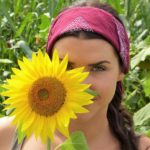A woman with a pink headband holding a yellow sunflower in a field.