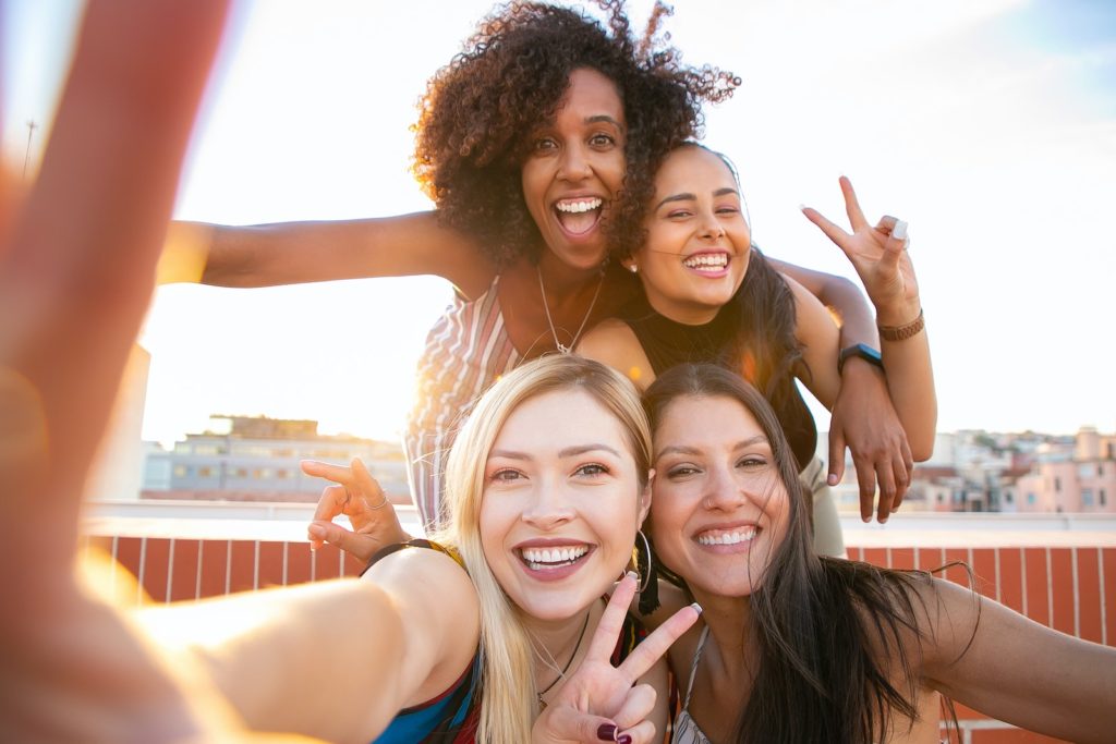 Cheerful young diverse women showing V sign while taking selfie on rooftop.Photo by Kampus Production.