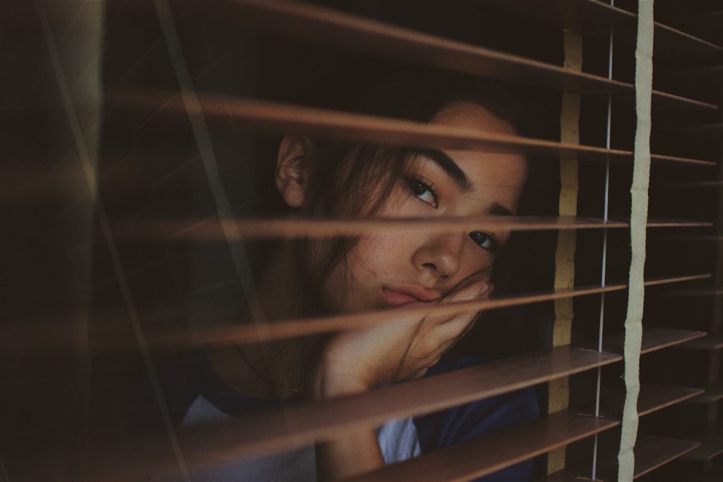 A woman looks through window blinds in a melancholy scene. Photo by Joshua Rawson-Harris.